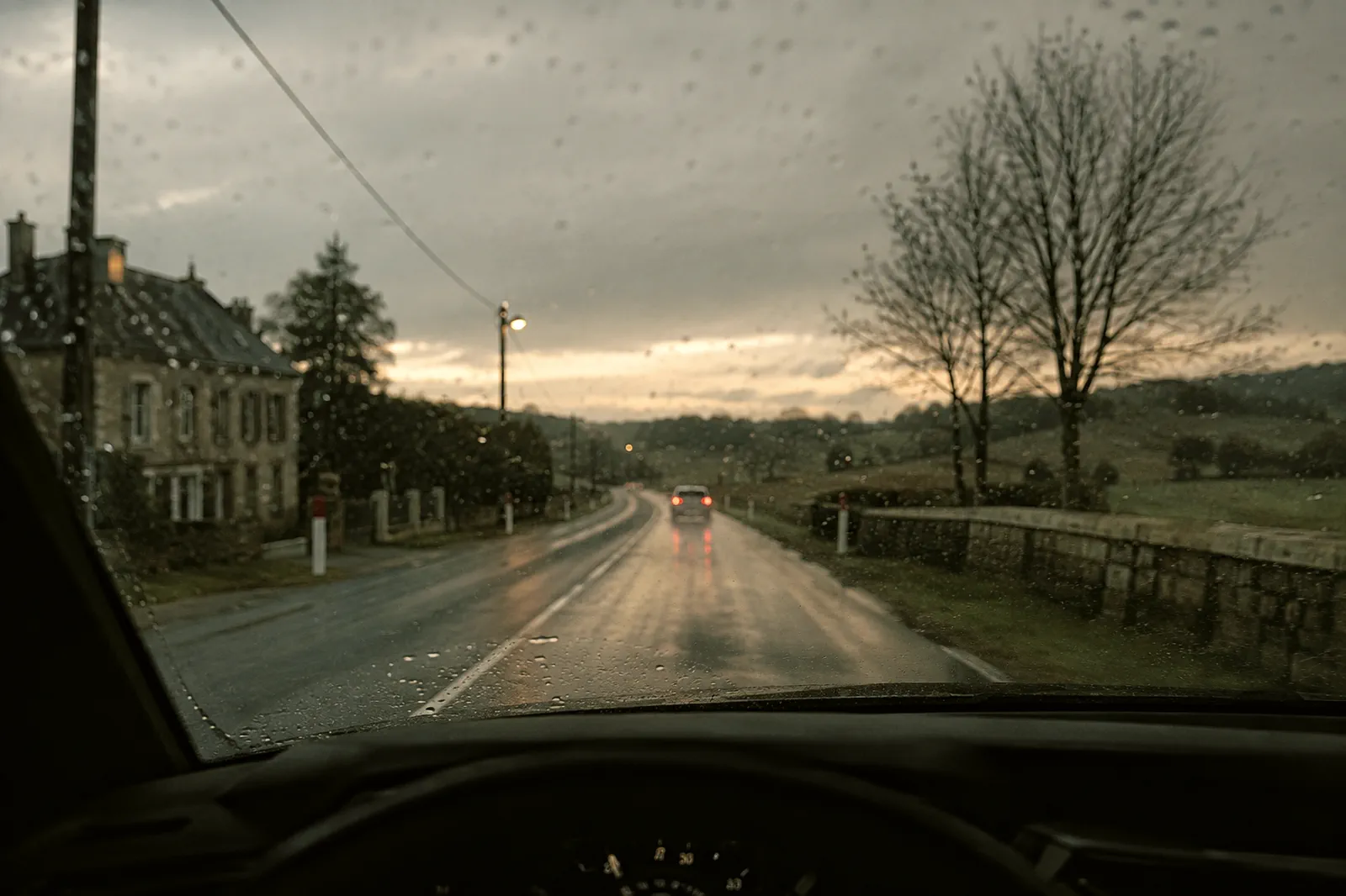 Route de campagne mouillée au crépuscule, vue depuis le siège conducteur, les feux d’un véhicule au loin.