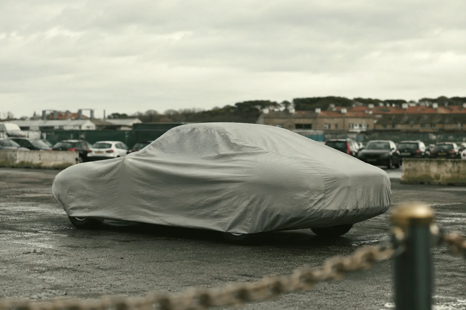 Un véhicule sous une housse de transport grise sur un parc d’importation, une chaîne au premier plan.